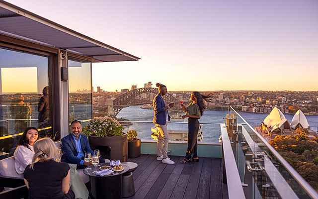 On the 32nd floor balcony of the Intercontinental Hotel in Sydney, a chic couple raises their glasses in a toast at Aster Bar as patrons enjoy a selection of foods on a table in the foreground. Bathed in the warm glow of the evening sun, they share smiles while sipping on delectable cocktails. The breathtaking harbour views provide a picturesque backdrop, enhancing the ambiance of this stylish and elevated setting.