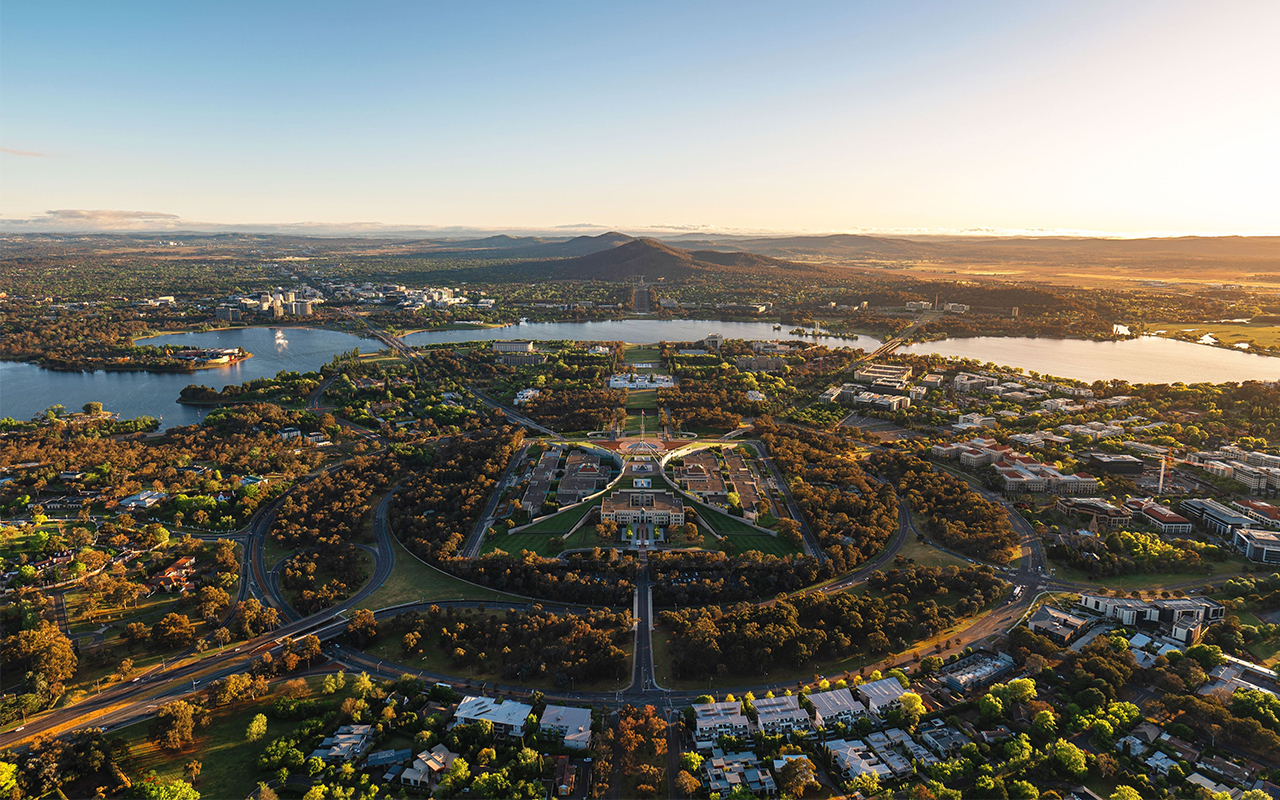 The aerial view of Canberra captures the iconic Parliament House prominently in the foreground. Beyond it, the older Old Parliament House can be seen. The tranquil waters of Lake Burley Griffin stretch out, reflecting the soft sunrise glow that bathes the city. In the distance, the solemn Australian War Memorial stands at the base of Mount Ainslie, which looms majestically in the background. The early morning light creates a serene and picturesque scene, highlighting the orderly layout and natural beauty of Australia's capital.