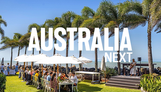 People dining at a long table under umbrellas Palm trees line the property with the ocean in the background. A singer stands on a stage nearby, entertaining guests © Tourism Australia