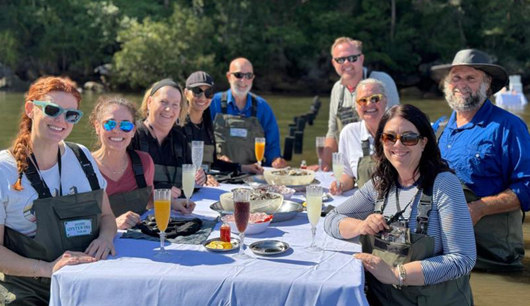 A group of North American agents standing around a table in water in waders, smiling for the photo. On the table are oysters and drinks in champagne glasses.