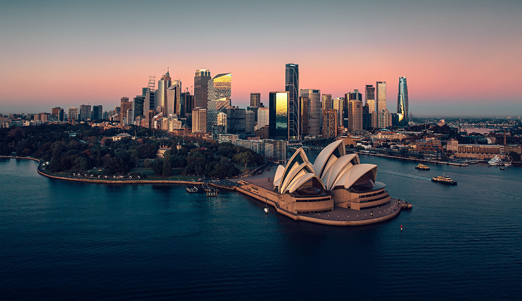 This captivating aerial image captures Sydney Harbour in all its splendor at sunset. The iconic Sydney Opera House gleams in the warm hues of the fading sun, while The Royal Botanical Gardens provide a lush green contrast. In the distance, Barangaroo's modern architecture harmonises with the city skyline, creating a stunning panorama of urban beauty against the backdrop of the tranquil harbour waters.