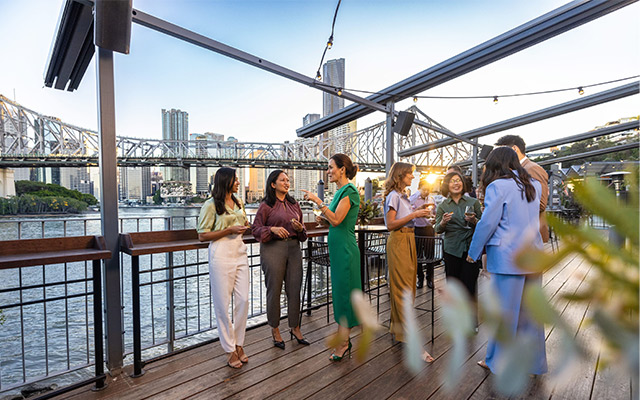 A group of people enjoy a relaxed evening by the Brisbane River, laughing and chatting as they savour cold drinks and canapés. They gather at Rivershed, where the warm hues of dusk paint the sky. Behind them, the Brisbane city skyline glows in the golden afternoon light, with the iconic Story Bridge prominently arching above, adding a touch of charm and character to the lively scene. The river reflects the city's glow, enhancing the festive yet tranquil atmosphere.