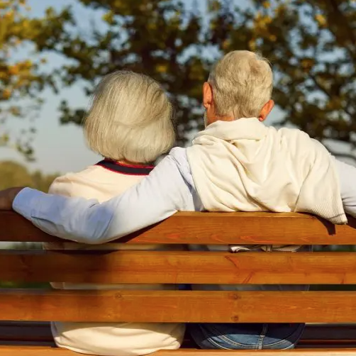 Two older adults sitting together on a park bench, arms around each other, looking out over a leafy outdoor setting.
