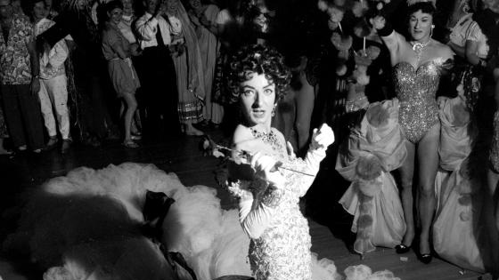 A black and white photo of a drag queen performing in a circle of people