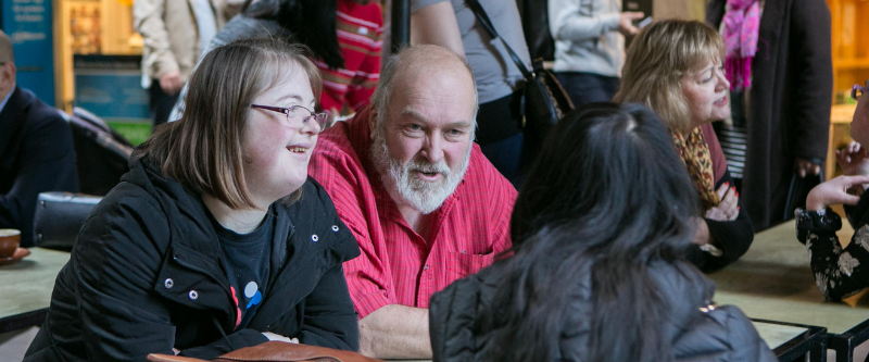 This is a photo of a female with Down syndome smiling. She is sitting next to a man and across from a woman at a table.