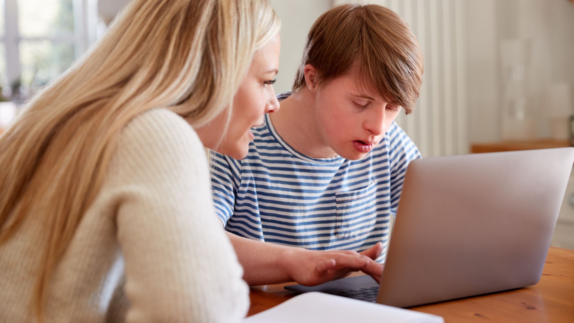 This is a photo of a teenager with Down syndrome on a laptop. His mum is sitting next to him.