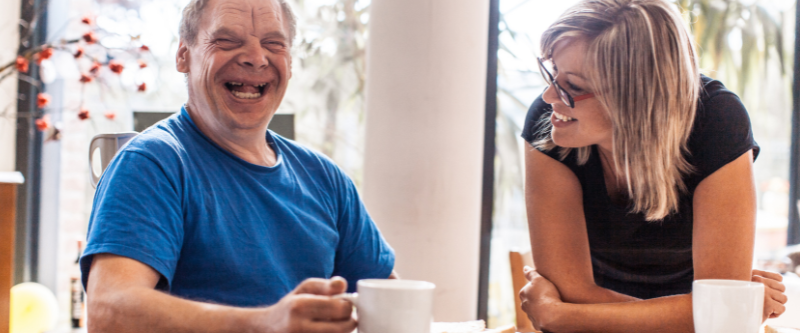 This is a photo of a man with Down syndrome drinking a cup of tea or coffee and smiling at the camera. He is with a women who may be his carer.