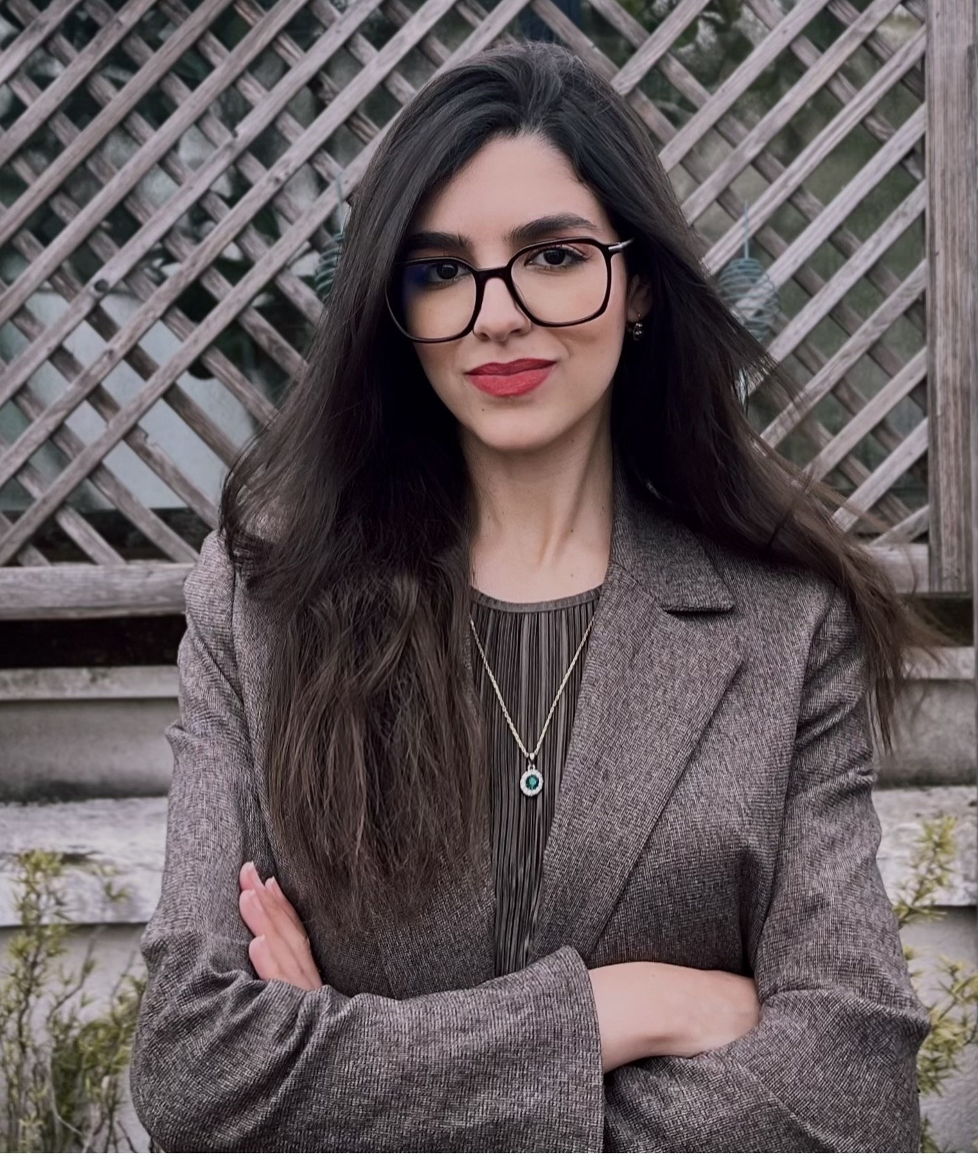 Sophia Hadef, of Keystone Procurement. She is standing in front of a garden fence with her arms crossed. She is wearing a jacket and a necklace.